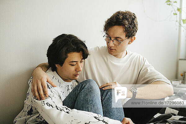 Young man sitting by worried male friend in bedroom