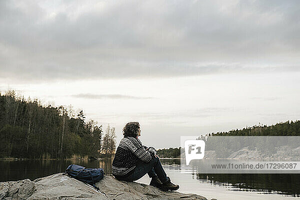 Mature female hiker spending leisure time while sitting on rock by lake during vacation