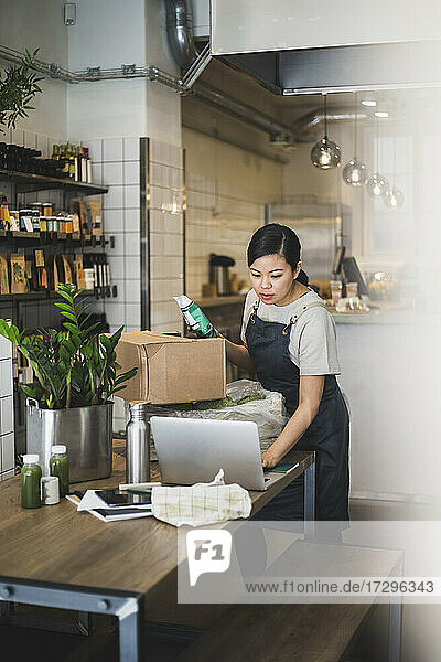 Weiblicher Besitzer arbeitet am Laptop  während er ein Lebensmittelpaket im Laden hält
