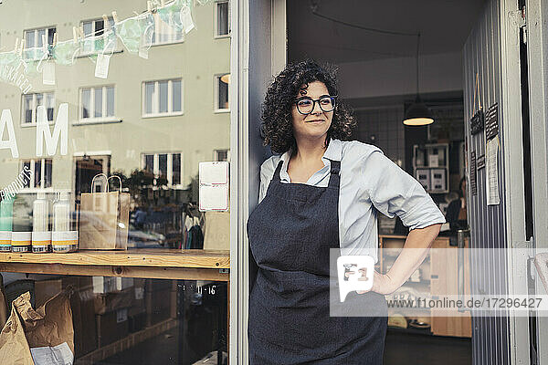 Happy female entrepreneur standing with hand on hip while looking away at store doorway