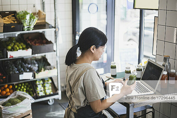 Asiatische Unternehmerin mit Laptop  während sie eine Saftflasche im Laden hält