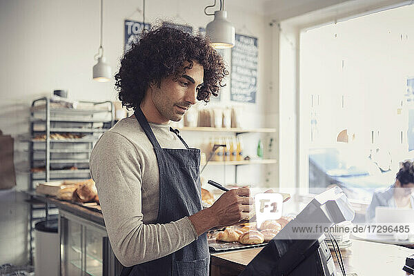 Männlicher Unternehmer schaut auf die Kasse  während er in einem Café auf einen Notizblock schreibt