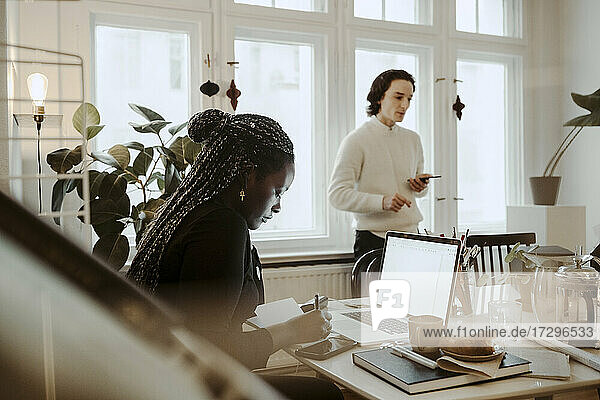 Weiblicher Profi schreibt am Schreibtisch  während ein männlicher Kollege mit Smartphone am Fenster im Home Office steht