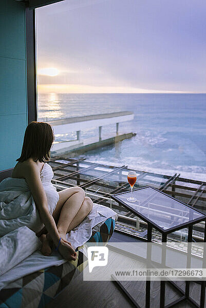 Frau sitzt auf dem Bett mit Cocktail im Hotel und beobachtet den Sonnenaufgang über dem Meer