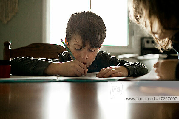 Junge macht Hausaufgaben mit einem Bleistift am Küchentisch mit Schwester