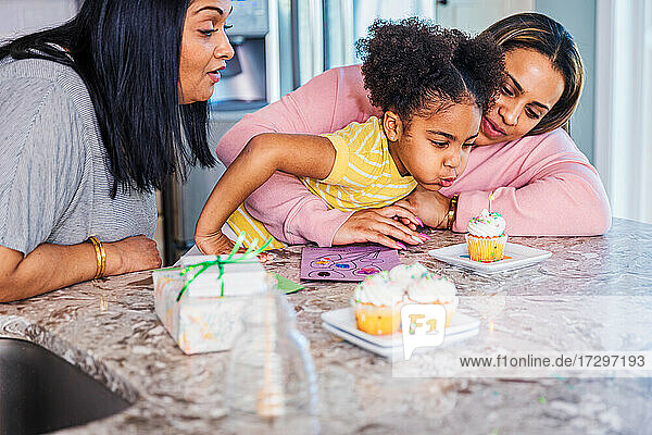 Mädchen bläst eine Kerze auf einem Kuchen aus  während sie mit Mutter und Großmutter zu Hause Geburtstag feiert