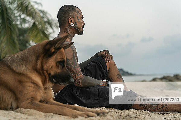Asiatischer Mann sitzt am Strand mit einem Hund unter Palmen