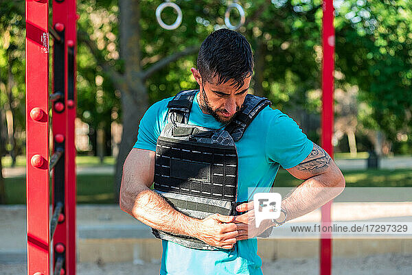 Dark-haired man with beard adjusting a weight vest in a fitness park. Outdoor fitness concept.