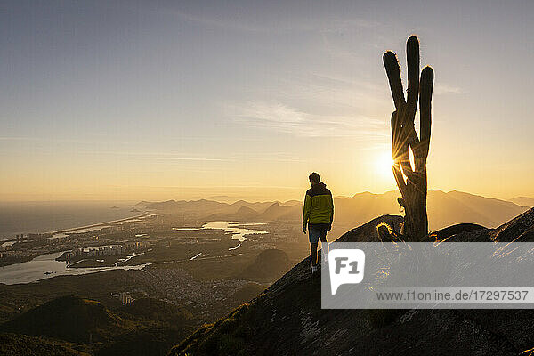 Man standing on rocky mountain edge next to big cactus on sunset