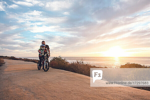 Father helping son ride his bike on a coastal trail at sunset.