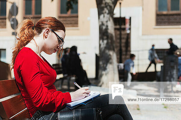 Junge rothaarige Frau mit Brille und roter Bluse sitzt auf einer Bank und schreibt etwas in ihr Notizbuch.