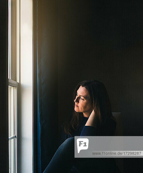 Woman sitting alone in a dark room looking out of the window.