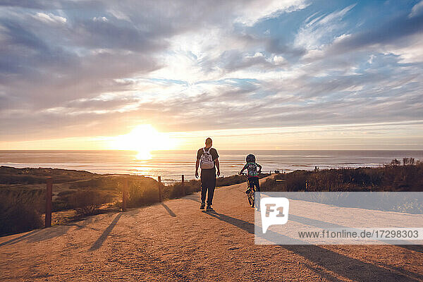 Father watching son ride his bike on a coastal trail at sunset.