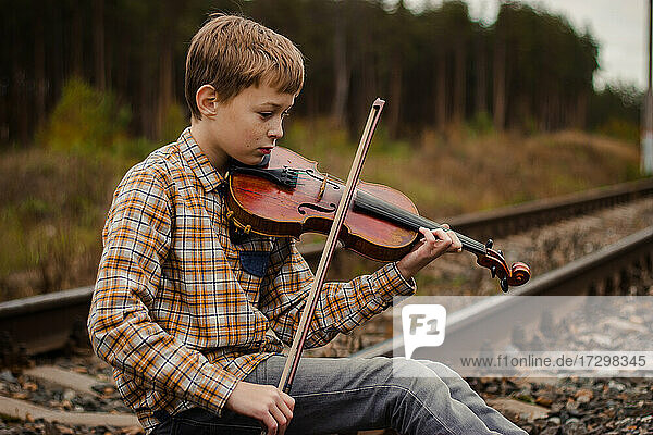 Ein schöner blonder Junge sitzt auf den Schienen der Eisenbahn und spielt Geige.
