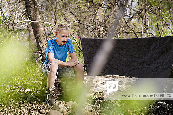 Junger blonder Junge sitzt bei seiner Festung im Wald.