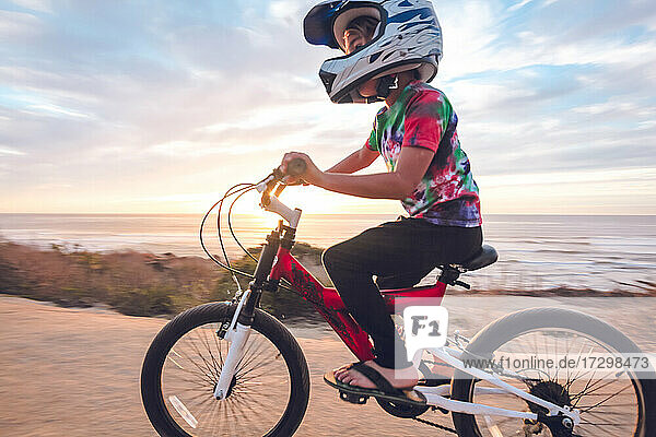 Kleiner Junge fährt mit dem Fahrrad auf einem Küstenweg bei Sonnenuntergang.