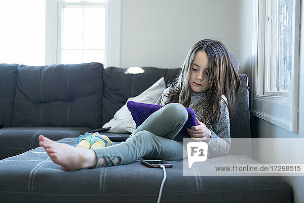 Kleines Mädchen sitzt auf der Couch und benutzt ein Tablet.