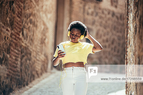 Porträt eines schwarzen Mädchens mit Afrofrisur  das mit Kopfhörern und ihrem Handy Musik hört  während es eine Straße in der Altstadt entlanggeht.