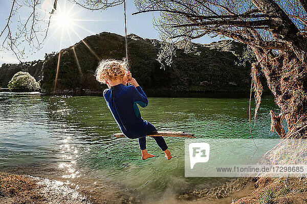 Blondes Kind auf einer Schaukel über einem Fluss in Neuseeland an einem sonnigen Tag