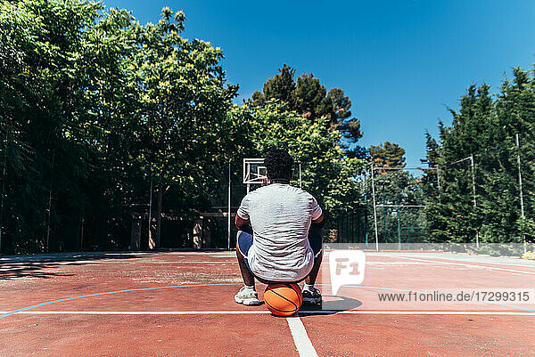 Rückansicht eines schwarzen Afro-Jungen  der auf seinem Ball auf dem Basketballplatz sitzt.