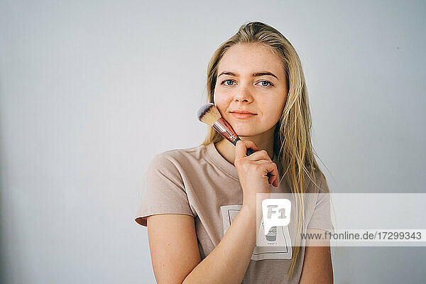 Blondes Mädchen mit einem Schminkpinsel auf grauem Hintergrund