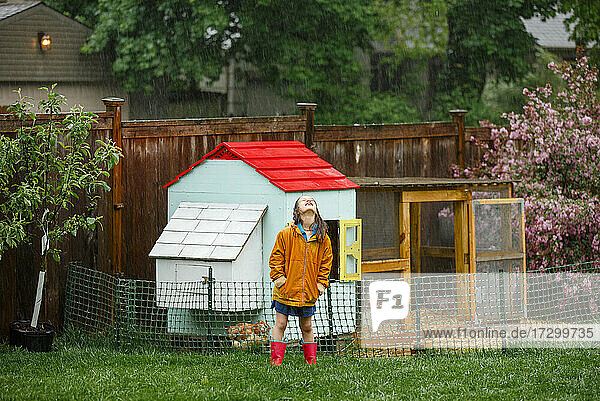 Ein fröhliches Kind steht im Regen im Garten beim Hühnerstall und schaut nach oben