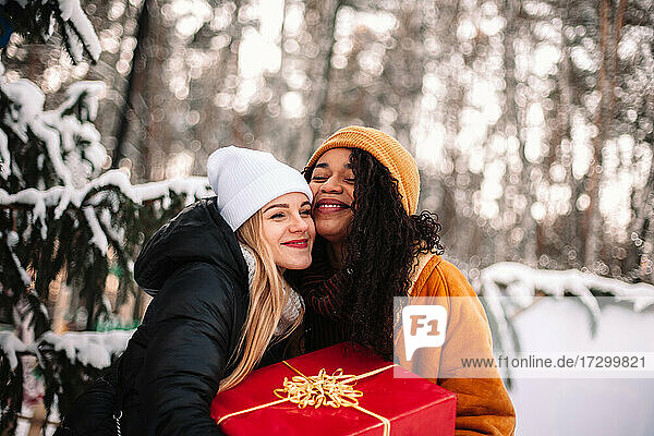 Happy girlfriends holding Christmas present standing by Christmas tree