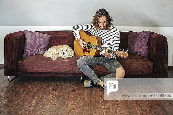 Attraktiver Mann mit langen Haaren spielt akustische Gitarre im Haus mit Hund