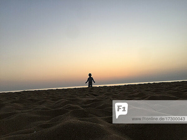 A little kid is standing on the beach with in sunset spreading hands
