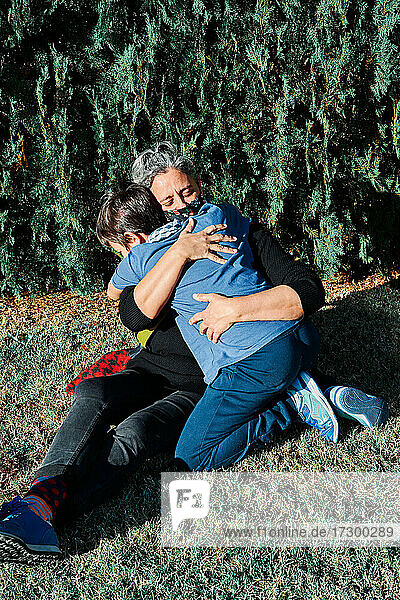 Vertical photo of a happy mother with a mask hugging her son sitting on the grass in the park. New normal