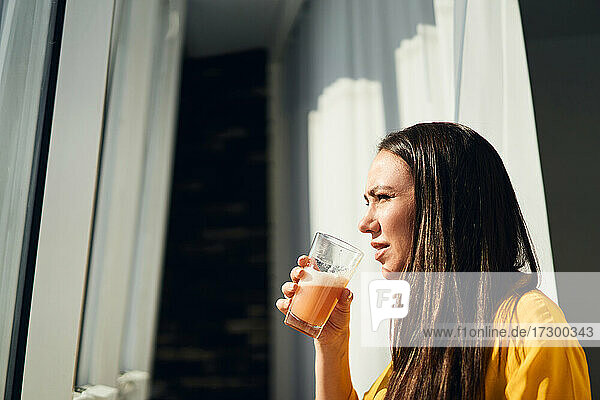 brünette Frau trinkt Saft aus einem Glas in der Nähe des Fensters