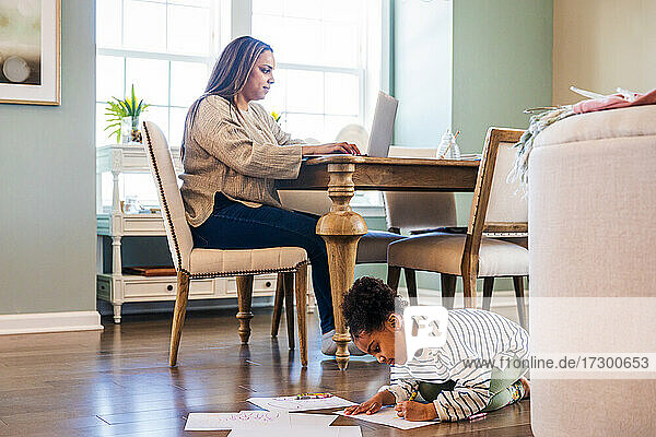 Niedliches Mädchen  das auf Papier zeichnet  während die Mutter einen Laptop benutzt  während sie am Tisch im Wohnzimmer sitzt