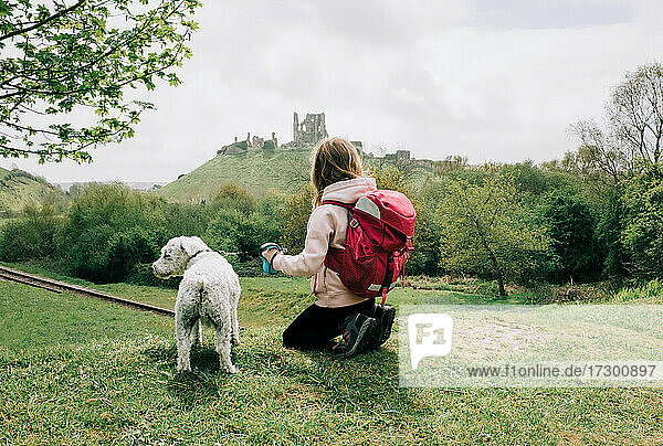 mädchen sitzt mit ihrem hund und schaut auf eine burg in der englischen landschaft