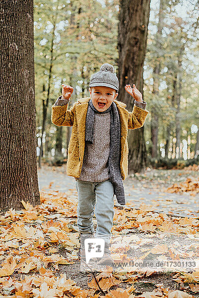 Stylish boy having fun in autumn city park. Happy kid walking among fallen leaves. Kids fashion. Boy wearing trendy yellow coat  cap and scarf. Smiling young boy outdoors. Kid jumping and run