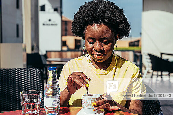 Porträt eines schwarzen Mädchens mit Afro-Haar und Ohrringen  das Kaffee und eine Flasche Wasser auf einer Bar-Terrasse trinkt.