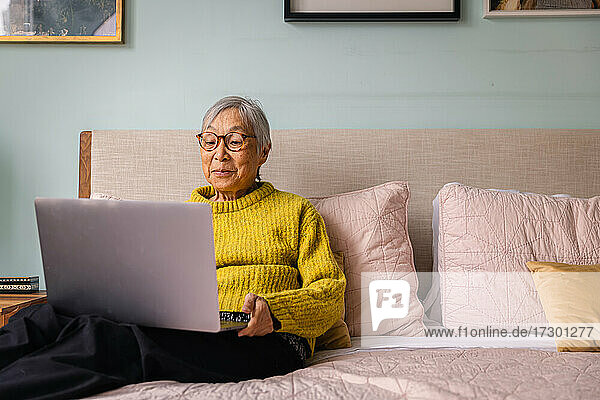 Ältere asiatische Frau  die einen Laptop benutzt  während sie im Schlafzimmer zu Hause sitzt