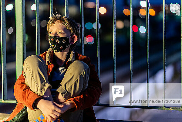 Teen with mask looking distant sitting against rail over train tracks