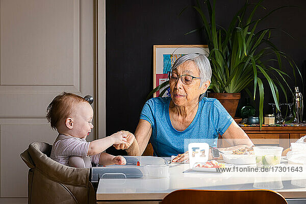 Senior asian woman feeding toddler grandaughter at table at home