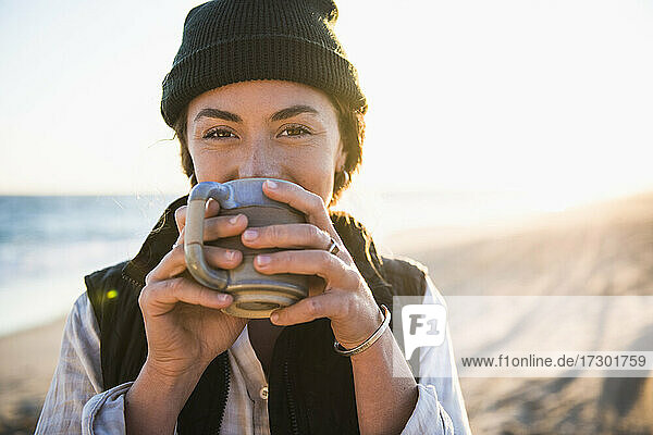 Junge Frau  die ein Getränk in einem Becher genießt  während sie allein im Auto am Strand campiert