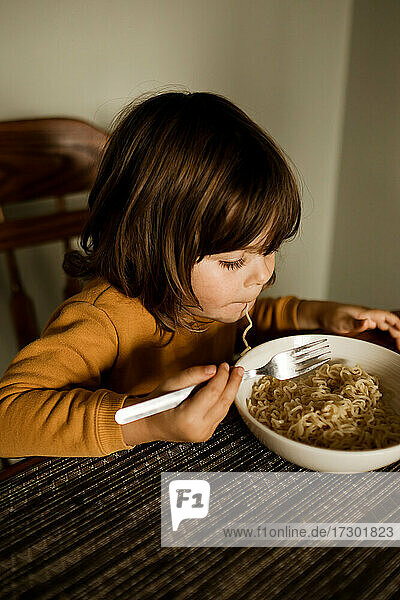 Junges Mädchen im gelben Pullover isst Nudeln am Küchentisch