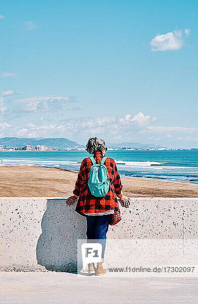 Vertikales Foto einer Frau mittleren Alters von hinten mit einem Rucksack  die an einem sonnigen Tag auf das Meer schaut