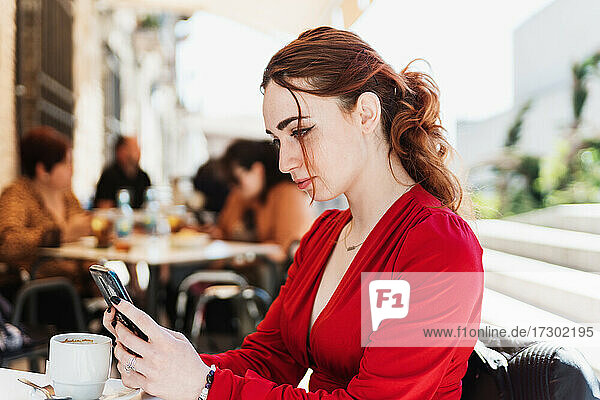 Junge rothaarige Frau in roter Bluse  die auf einer Terrasse Kaffee trinkt und dabei ihr Mobiltelefon benutzt.