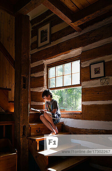 Ein Junge liest ein Buch auf der sonnigen Treppe eines Blockhaushauses.