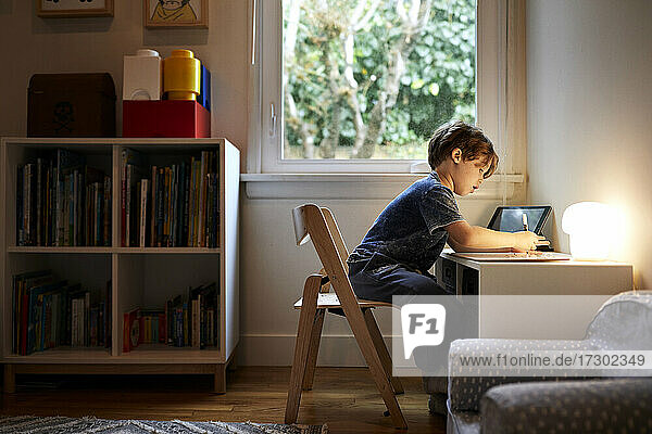 Seitenansicht eines Jungen  der zu Hause auf einem Stuhl am Fenster sitzt und zeichnet