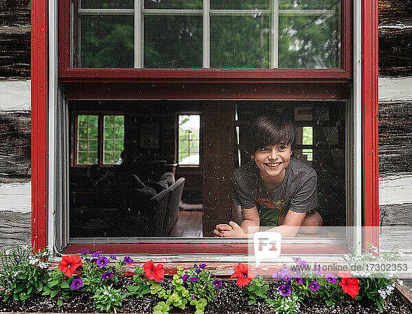 Junger glücklicher Junge schaut an einem Sommertag aus dem offenen Fenster einer Blockhütte.