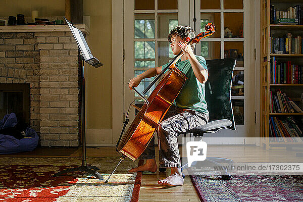 Ein barfüßiger Junge übt Cello im goldenen Fensterlicht im Haus