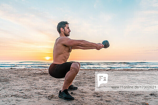 muscular man training legs with kettlebell on the beach at sunrise