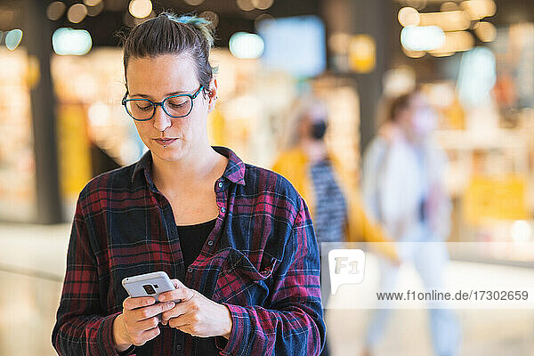 Spanische Frau tippt zweifelnd auf ihrem Smartphone  während sie in einem Einkaufszentrum einkauft
