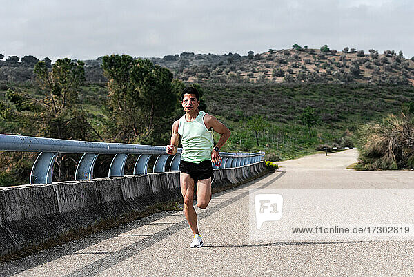Junger lateinamerikanischer Läufer  der auf einer Brücke läuft. Straße und Wald im Hintergrund.