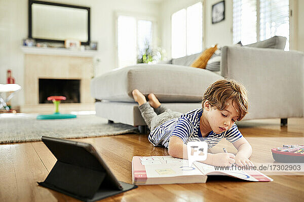 Boy lying on hardwood floor while coloring in book with crayon at home
